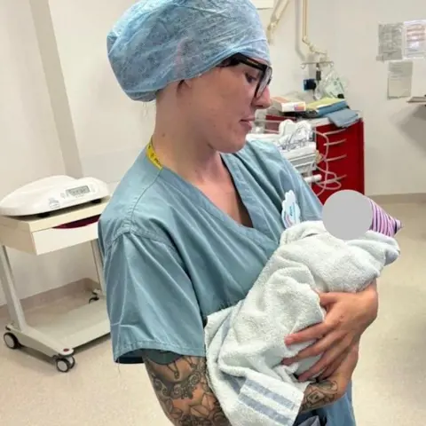 Supplied A woman in blue scrubs is holding a baby in a room in a hospital. The baby is wrapped in a white towel with a pink and white hat on its head. It's face has been obscured. Medical equipment can be seen in the background.