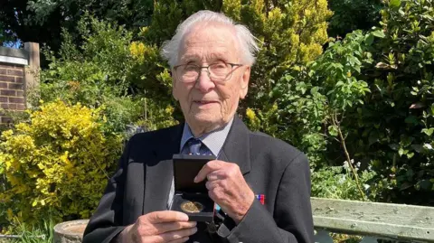 Ken Foster in a suit holding a medal with other medals pinned to his chest