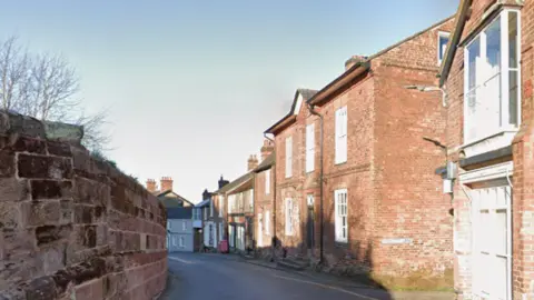 Google A narrow street in a village, lined by a wall on one side and terraced housing on the other.