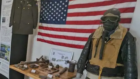 Stanwick Lakes A large US flag behind a wooden shelf of artefacts, mainly rusty plane parts. A mannequin has been placed next to the shelf wearing World War Two flying gear including a hood, goggles, life jacket and breathing tube.