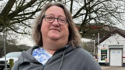 Clare Worden/BBC A woman with shoulder-length brown hair wearing glasses. She is wearing a blue and white floral patterned top and a grey zip-up hoodie. She is looking at the camera and smiling