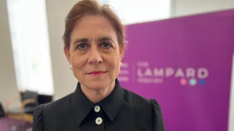 A woman with dark brown hair which has been tied up is looking at the camera. She is wearing a black blouse with cream buttons. A large purple banner is behind her with the word 'Lampard' visible.