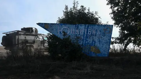 A Ukrainian military vehicle drives past a welcome sign to the Yunakivka village in Sumy, Ukraine, which is a checkpoint through the state border of Ukraine on the border with Russia. The sign is triangular and blue with white writing in the centre of the image. The vehicle is to the left of the sign. there are trees surrounding it.