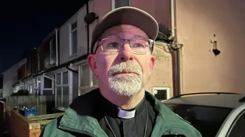 BBC/Amanda White Reverend Ian Greenfield is wearing a baseball cap, a shirt with a dog collar and a dark green coat. He has a short grey beard.