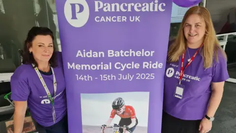 Supplied Two women dressed in purple T-shirts stand either side of a large purple banner with the words "Memorial Cycle Ride".