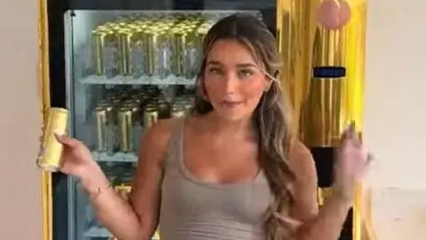 PA Media Lucinda Strafford  standing in front of a vending machine. the machine is gold with a window in front showing it is full of gold cans of a u vodka. She is holding a can up in one hand. she is a young looking woman with a crop top, make up and pull back long blonde hair. She is holding her other had up and is smiling. 