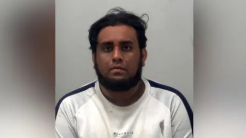 A man wearing a white top with a black strip down the shoulder. He has a beard and he is looking at the camera. It is a custody photo.