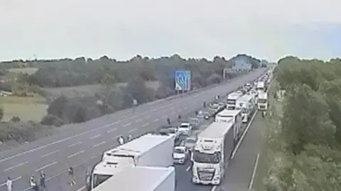 National Highways A motorway camera view showing a queue on one carriageway, People can be seen out of their cars. 