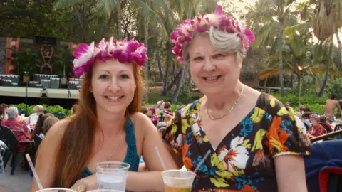 Elaine Murray A photo of Elaine and her mum. Elaine is wearing flowers in her hair which are purple. So is her mum. Her mum is wearing a floral top and a necklace.