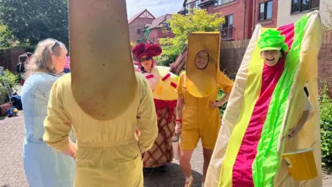 People dressed up as food, including chips and a sandwich, wait around in a circle