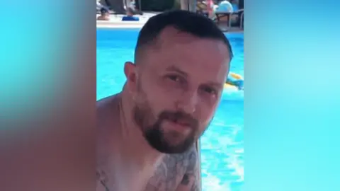 A photo of Stephen Holmes. He is shown from the shoulders up sitting in front of a pool. He has short brown hair, and a beard. He is smiling at the camera.