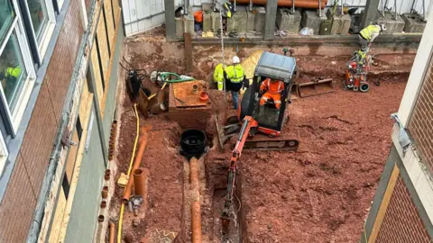 An overhead image of construction workers at work. The ground is brown and there is machinery around. One worker is in a digger which is digging a hole in the ground. Behind it are three mean in green high visibility clothing. To the left is a wall and windows. 