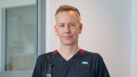 A nurse stands beside a window and a white wall. He wears a black nurse's overall. NHS is branded on this chest
