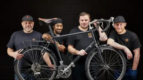 ONWARDS AND UPWARDS Four men, all wearing black t-shirts, stand behind a racing bike and in front of a black background.