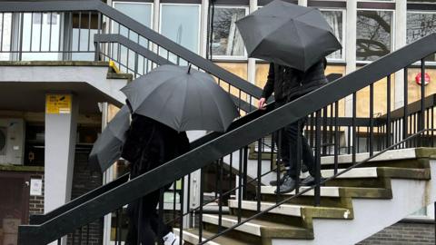 Image of Sophie Harvey and Elliot Benham outside Gloucester Crown Court. They are pictured under black umbrellas concealing their faces. 