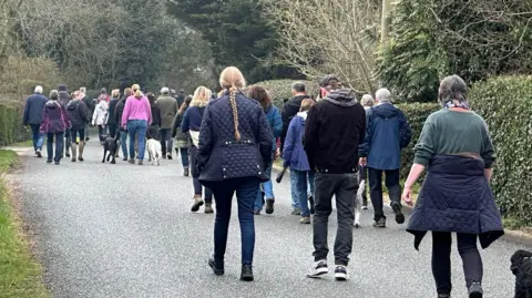 A crowd of people walking down a country road with greenery either side of them