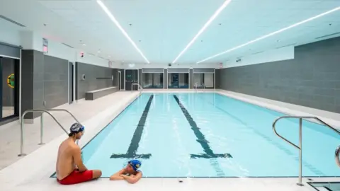 Two people sitting at the side of the pool. One inside the pool. Both wearing swimming caps 