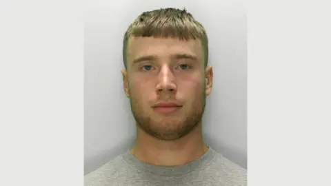 Gloucestershire Police A police mugshot of Kane Pearson. He is wearing a grey T-shirt and is standing against a light grey background. He has dark blonde hair which is shaven on the sides and cut into a straight fringe on top. He has blue eyes and a dark blonde beard and is looking directly at the camera.