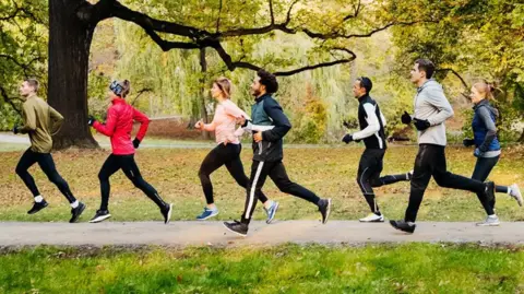Seven people, men and women, are dressed in a variety of running gear and pictured running through a park on a tarmac path in front of trees.