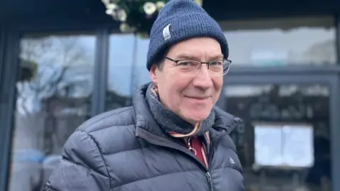 Elizabeth Baines/BBC A man in a puffer jacket and navy beanie hat with glasses and a grey scarf.