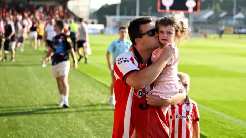 Stephen McCarthy/Getty Images Shows a man wearing a Derry Citystrip carrying a young girl who is crying. Other fans are in the background.