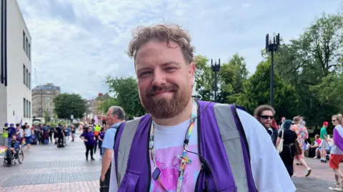 Ste Dunn director of Northern Pride being interviewed. He is wearing a purple high viz jacket and is standing in front of the Pride parade. He has short, curly blond hair and a reddish beard.