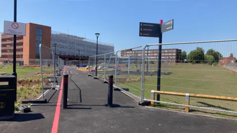 A view of works on the Royal Arcade site to create a park. A path is in the middle with fenced off grass areas and a play park in the background.
