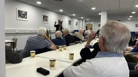 George Carden/BBC people sitting in a white room looking at a musician playing cover songs, people have coffees on the table 