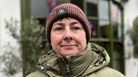 Davina Love is wearing a green puffer coat, a brown hat and is smiling. She has several nose piercings.