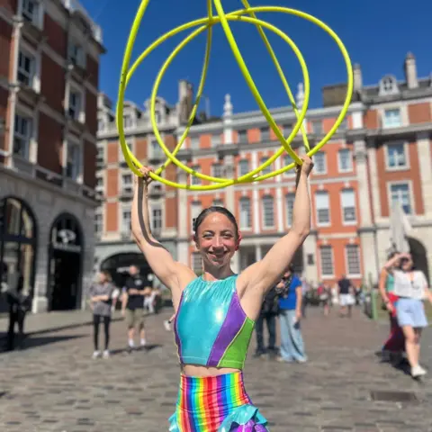 A woman in a rainbow-coloured outfit, holding a combination of bright yellow hoops above her head.