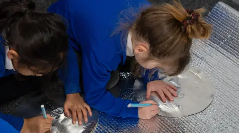 Two girls in blue uniforms are focused on etching designs into metal plates
