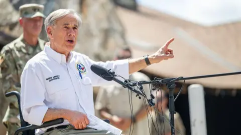 Texas Governor Greg Abbott, who uses a wheelchair, is seen seated before a microphone as he speaks and gestures. He is wearing a white shirt and khaki pants. There is a man in military fatigues slightly out of focus behind him. 