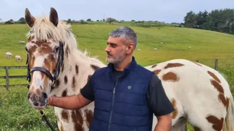 Supplied Saundh is holding the reins of white horse with brown spots. He is wearing a black t-shirt and a blue body warmer. They are on a field with sheep grazing in the background. 