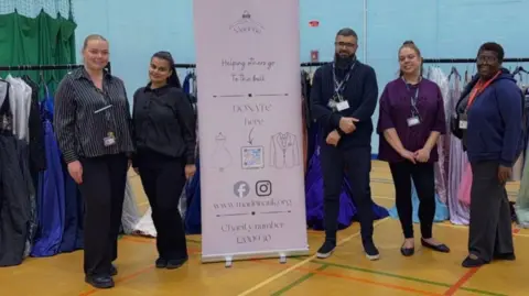 Erdington Academy A group of people stand beside a display sign in a sports hall, with rails of formal dresses arranged behind them.
