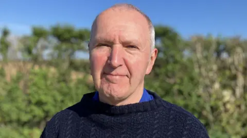 Man with a slight smile on his face. He has a bald head and is wearing a blue linen shirt with a fisherman type sweater on top. 