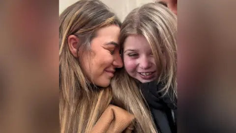 Family photograph Side profile of Francesca (left) with long brown hair wearing a tan jacket with her eyes closed and face resting on the cheek of her daughter, Sienna, who also has long brown hair and is wearing a black scarf. Sienna is smiling.