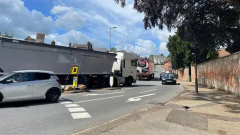 A road with cars and trucks driving along it with hedges to the right and a house further up the hill in the background.