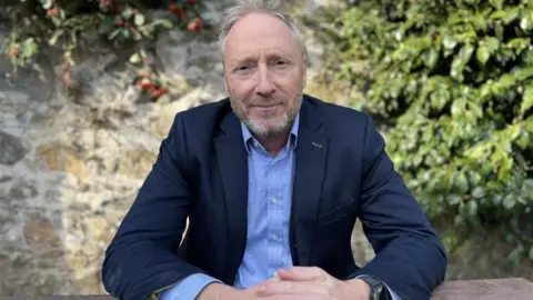 Deputy Paul Montague - a man with grey hair, sitting down with his arms in front of him, hands clasped together. He's wearing a blue suit with a blue button up shirt.