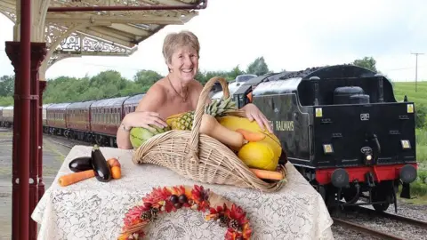 Nikita Kayleigh Photography An older woman with short light brown hair sitting in front of a basket of fruit and vegetables, including a melon, carrot and butternut squash. A train is in the background. An aubergine and carrot are on the table next to her.