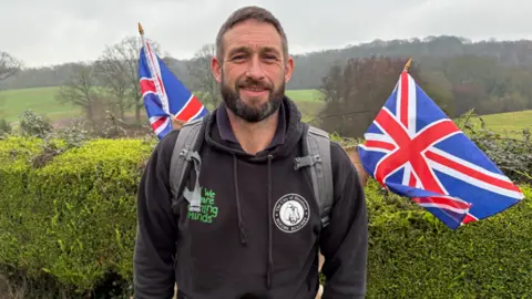A man with dark hair and beard wearing a black hoodie with a green We are Farming Minds logo. He is standing outside in front of a hedge, with fields visible behind him and two Union Jack flags sticking out of a rucksack on his back.