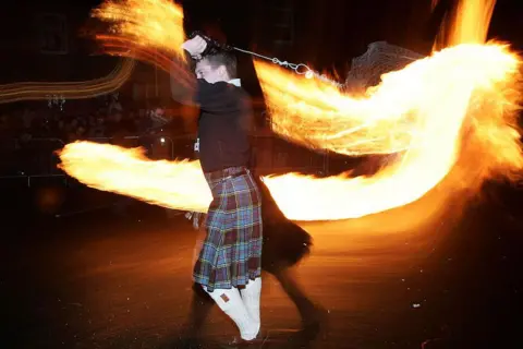 Getty Images A man in a kilt swinging a fireball on a chain around his head. The photograph shows the trail of fire 