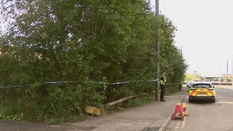 Wide shot of two police officers standing on a pavement in front of a wooded area that has been cordoned off with blue and white police tape. There is a police car among vehicles parked on the road in front of them.