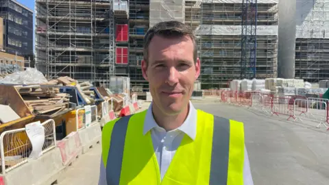 Ben Schofield/BBC Matthew Pennycook with short brown hair wearing a bright yellow high-visibility vest stands at the front of an active construction site. Behind the person, a multi-storey building is covered in scaffolding and safety netting, with materials, barriers, and construction equipment arranged along a partially completed road. The scene is brightly lit by sunlight, showing details of the building works and surrounding structures.