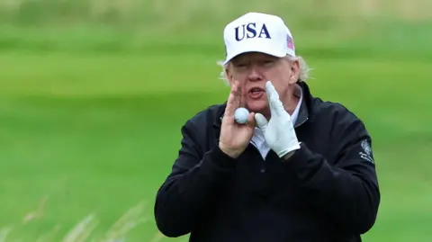 Reuters Trump holds his hands to his mouth and shouts while on the golf cours. He is holding a golf ball in his right hand and is wearing a black waterproof jacket, white glove and white hat with USA on the front