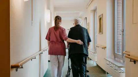 Nurse wearing red top and white trousers supporting elderly person walking down a care home hall. It has a green carpet and white walls.