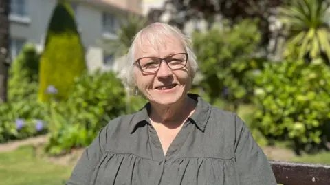 Jamie Poindexter is sat on a bench in a garden. She is wearing a dark green shirt and glasses and smiling into the camera.