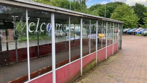A large roofed bus shelter with glass panelling and a red rim around the bottom, with the words "Welcome to Oxon Park and Ride" stencilled onto the windows. There is herringbone paving around it, and cars parked under trees in the background.