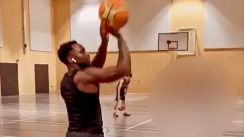 Supplied Benjamin Katabana on a court playing basketball. He has airpods in his ears and is wearing a black top. There is another man playing basketball in the distance. 