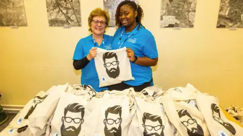 NWAFT Two women holding a cloth bag with a male face sporting a beard and moustache. They are surrounded by bags with the same print on a table.