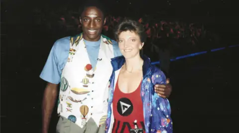 Nicola Bawden on the right and John Fashanu on the left with their arms around each other. Bawden is wearing the gladiators attire and a colourful jacket and Fashanu is wearing a blue t-shirt and a waist coat with a variety of images of hot air balloons on.